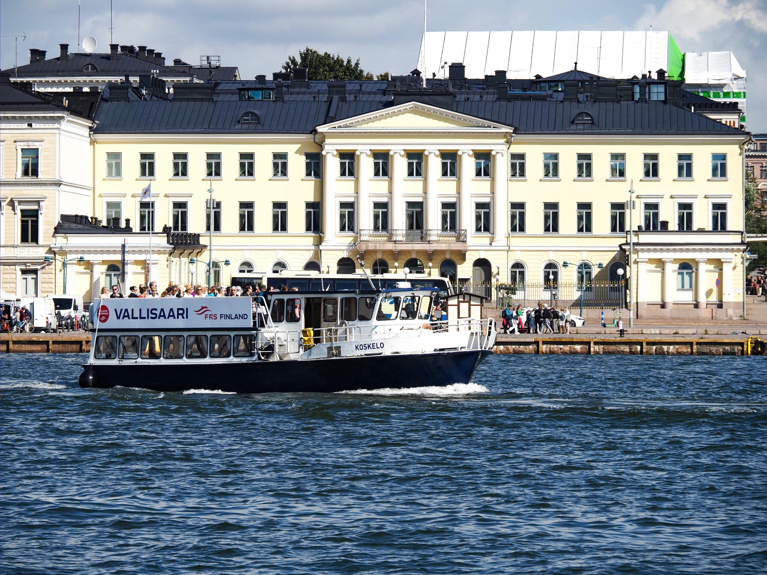 A boat that is en route to Vallisaari from the Market Square. In the background, a large light-colored building can be seen.