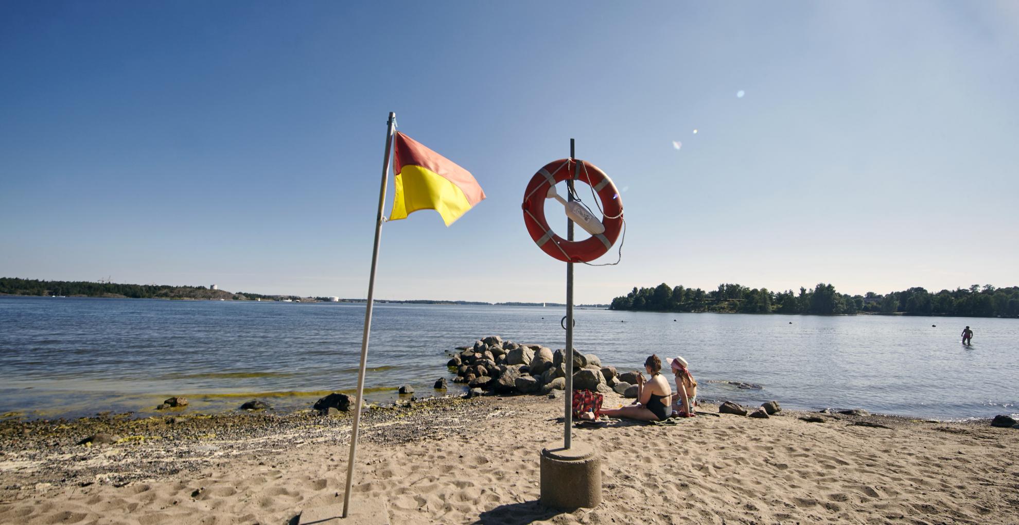 Henkilöitä Mustikkamaan rannalla. Liehuva lippu, ja taustalla merihorisontti.